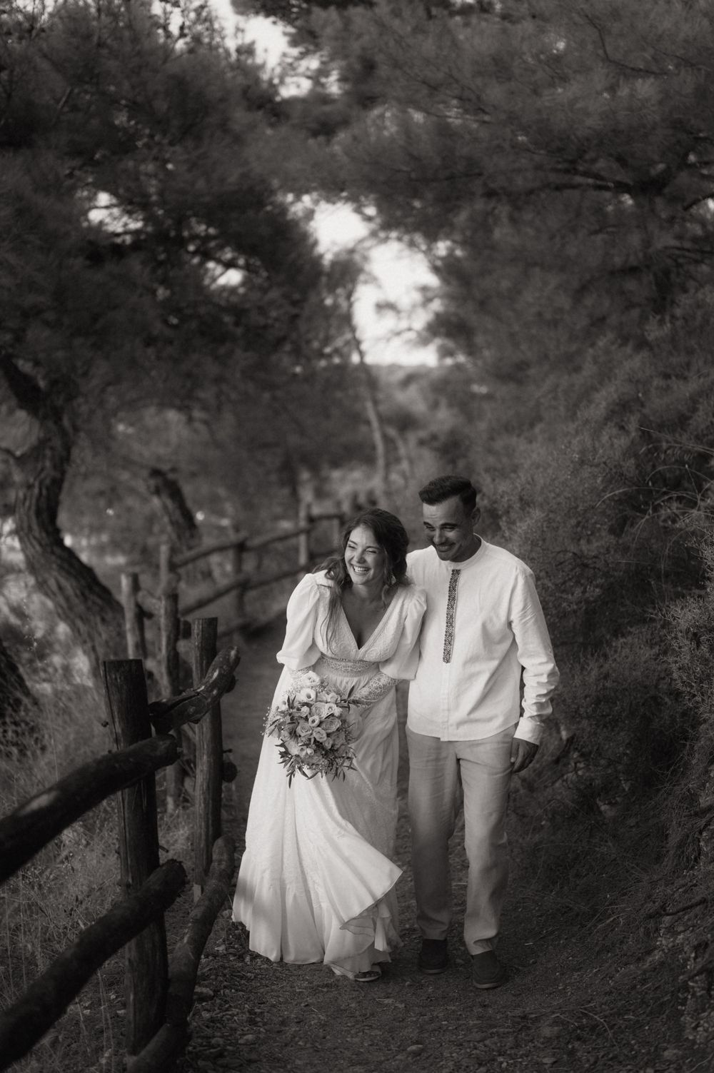 Black and white photo of a smiling bride and groom walking a tree-lined path, bride holding a bouquet.
