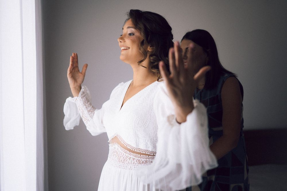 Bride in white lace dress smiling near window while bridesmaid helps her prepare for wedding day.