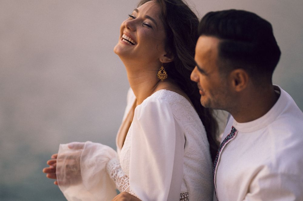A joyful couple in white outfits laughing together outdoors, sharing a happy romantic moment.