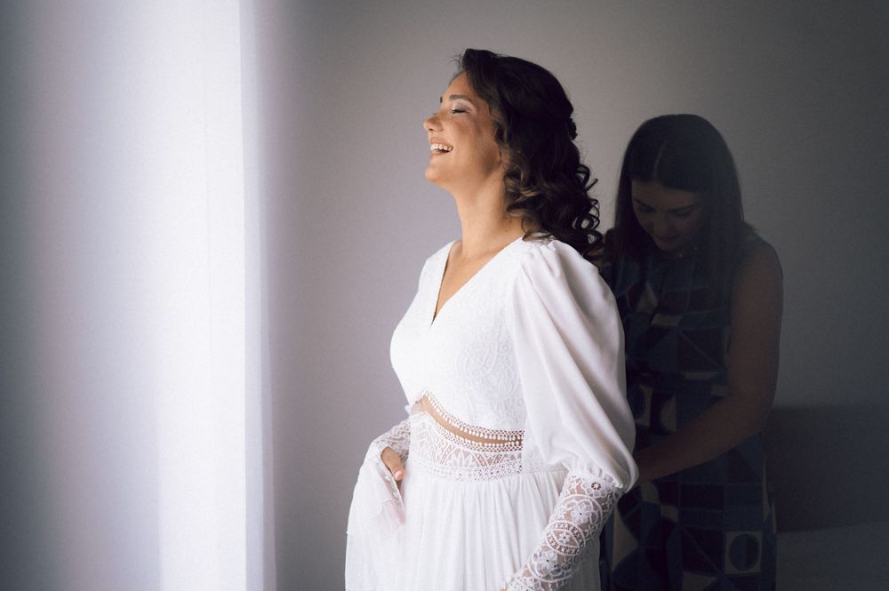 Smiling bride in white wedding dress being helped by bridesmaid near bright window.