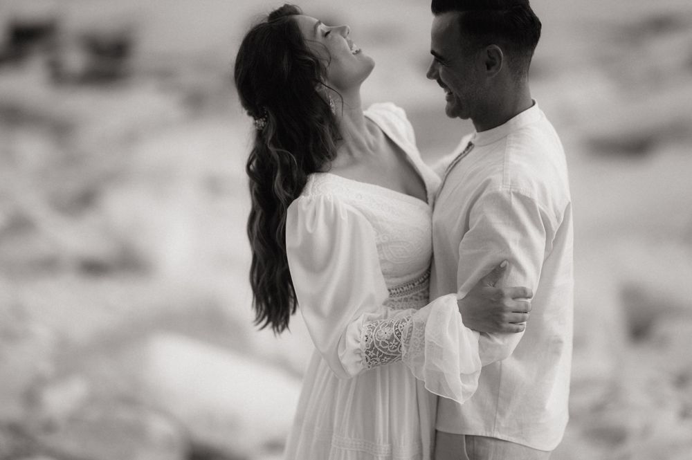 Black and white photo of a joyful couple embracing outdoors, woman in white dress laughing, man in white shirt smiling.