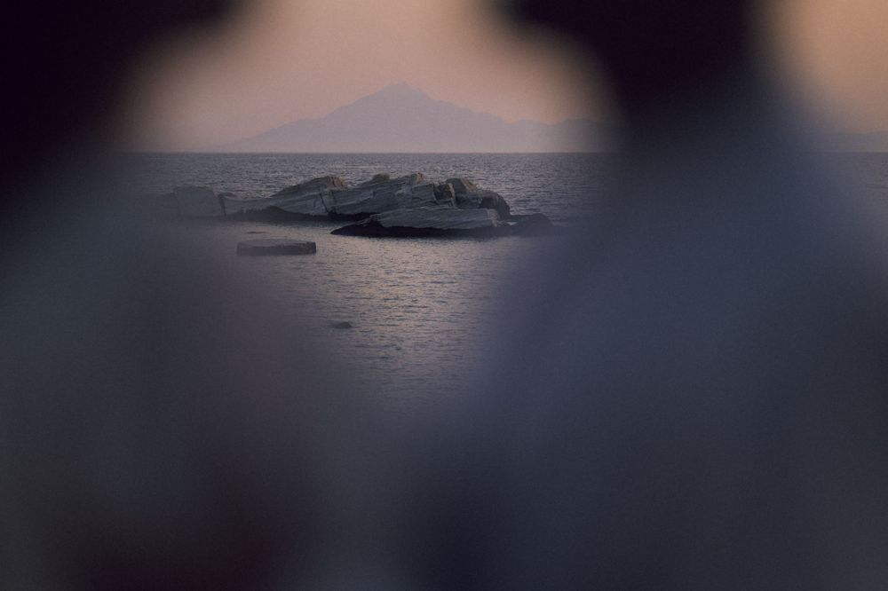 Misty seascape at dusk with rocky outcrop and distant mountain silhouette framed by blurred foreground.