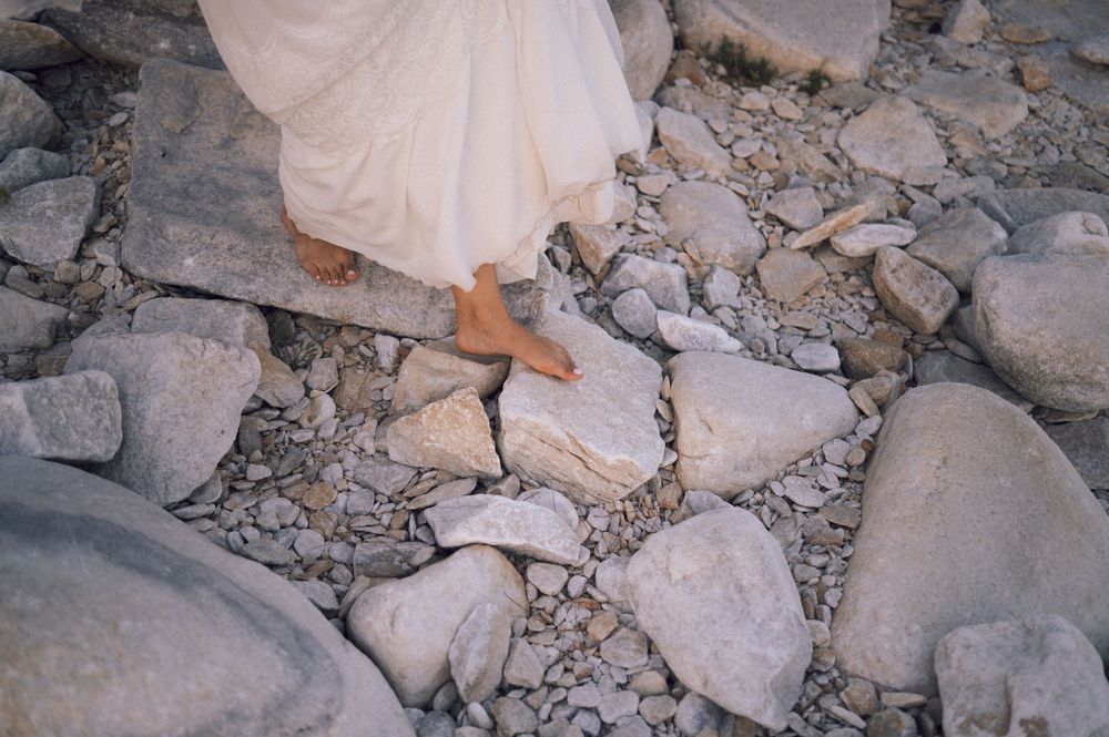 Barefoot person in white dress walking carefully over rocky terrain with large stones and pebbles.