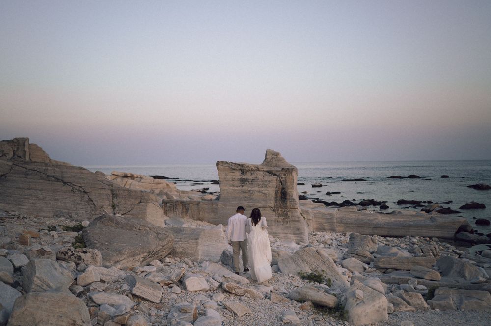 Couple standing among ancient coastal ruins at dusk with rocky shoreline and pastel sky backdrop.