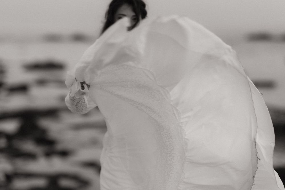 Black and white photo of a woman in a flowing white dress billowing in the wind by the ocean shore.
