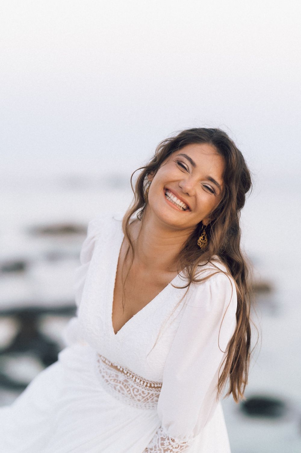 Smiling young woman with long brown hair wearing a white lace dress and gold earrings posing near the ocean.