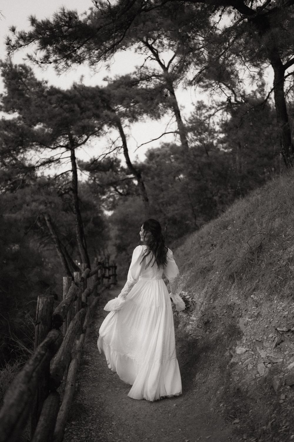 Black and white photo of a bride in a flowing white gown walking along a forest path holding a bouquet.