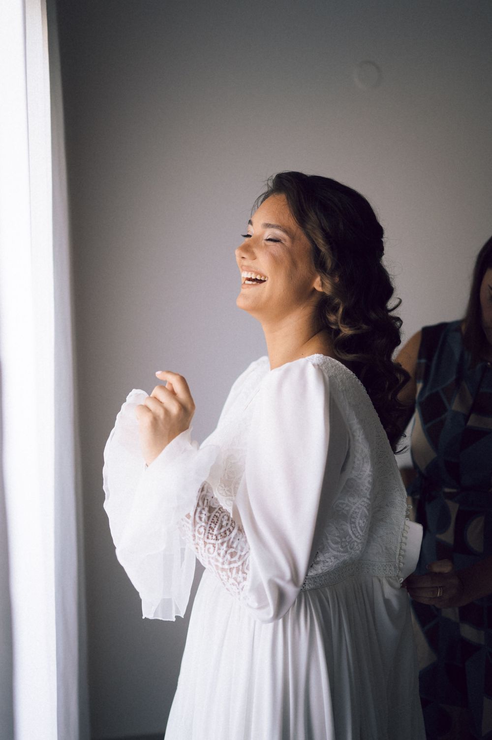 Joyful bride laughing in white lace wedding dress standing by bright window.