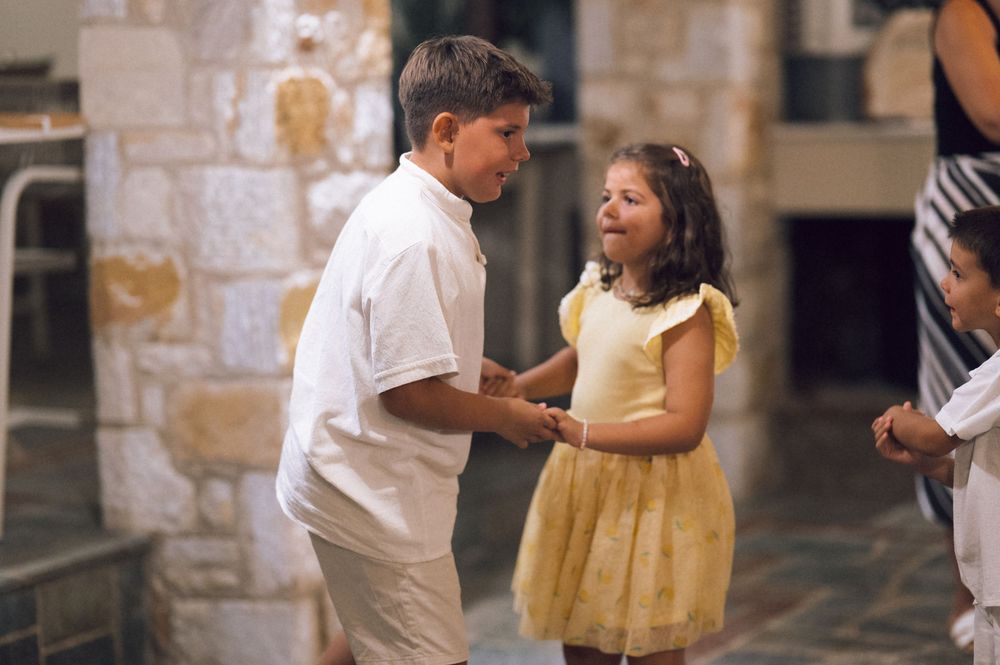 Two children, a boy in white and a girl in yellow dress, interact outdoors near stone architecture.