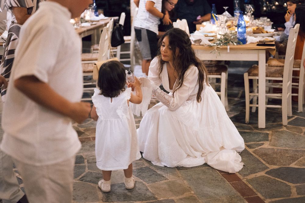Bride in white gown crouches to interact with young girl in white dress at an outdoor wedding reception.