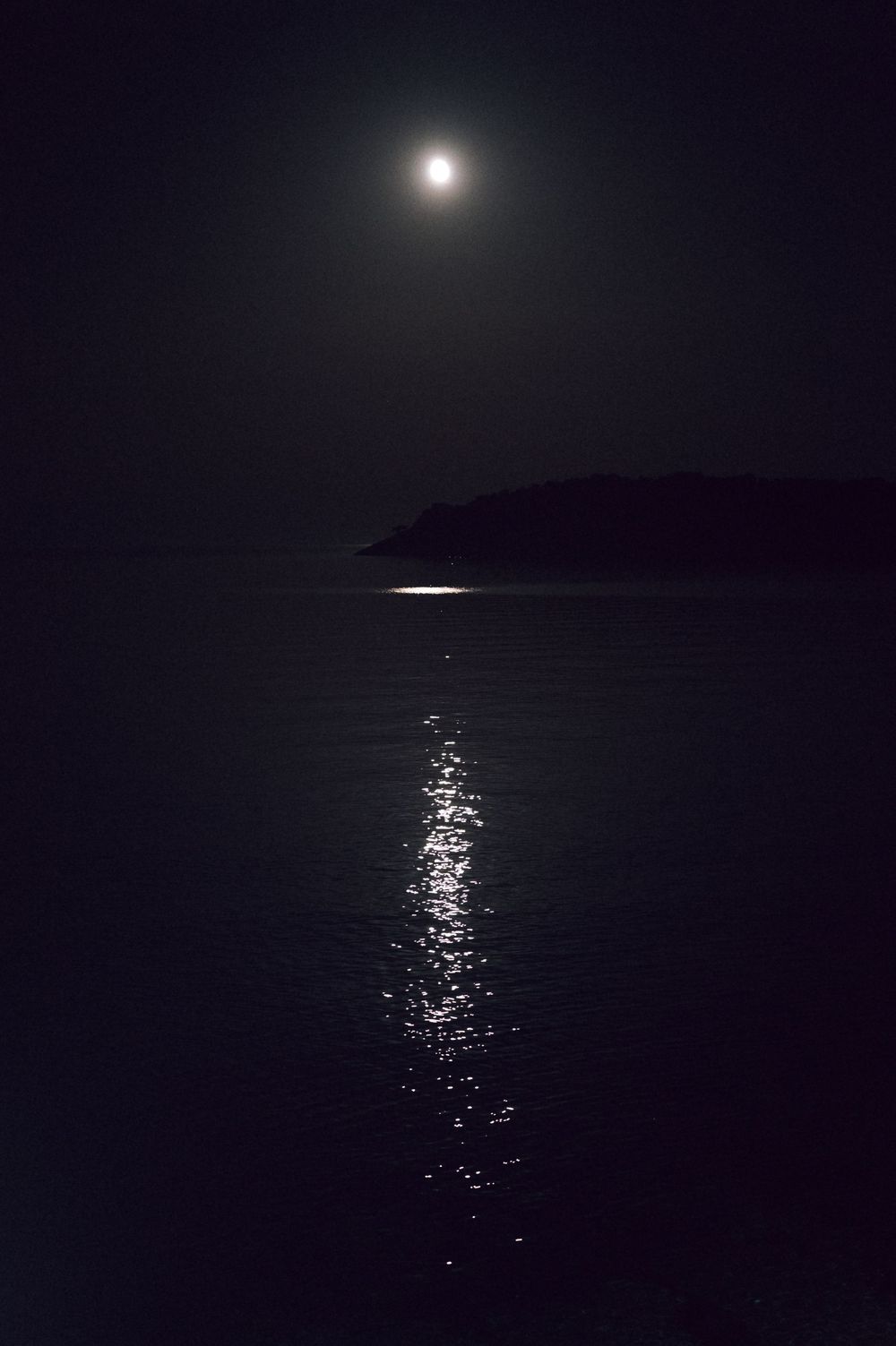 Moonlit night seascape with glowing reflection on calm ocean water and dark silhouetted island in background.