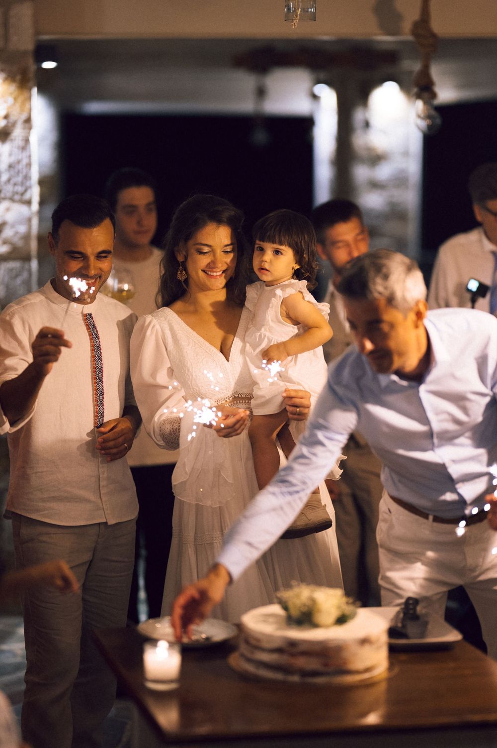 Wedding couple with sparklers cutting cake, bride holding child in white dress at elegant evening reception.