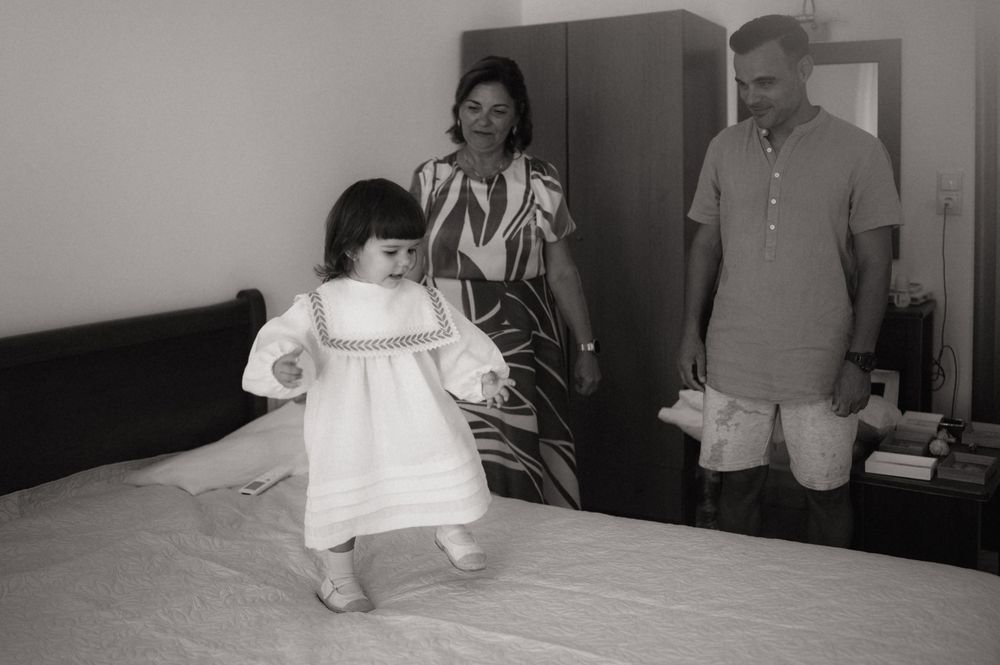 A young girl in a white dress stands on a bed while her parents watch in a black and white family photo.