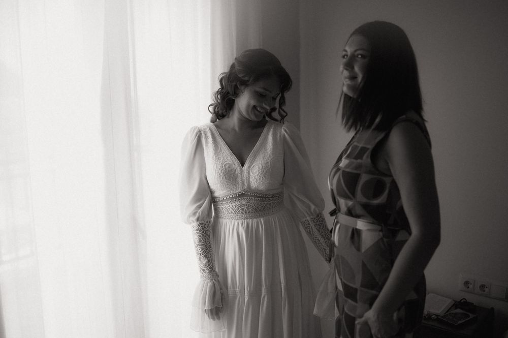 Black and white photo of a bride in white gown sharing a tender moment with a bridesmaid near a window.