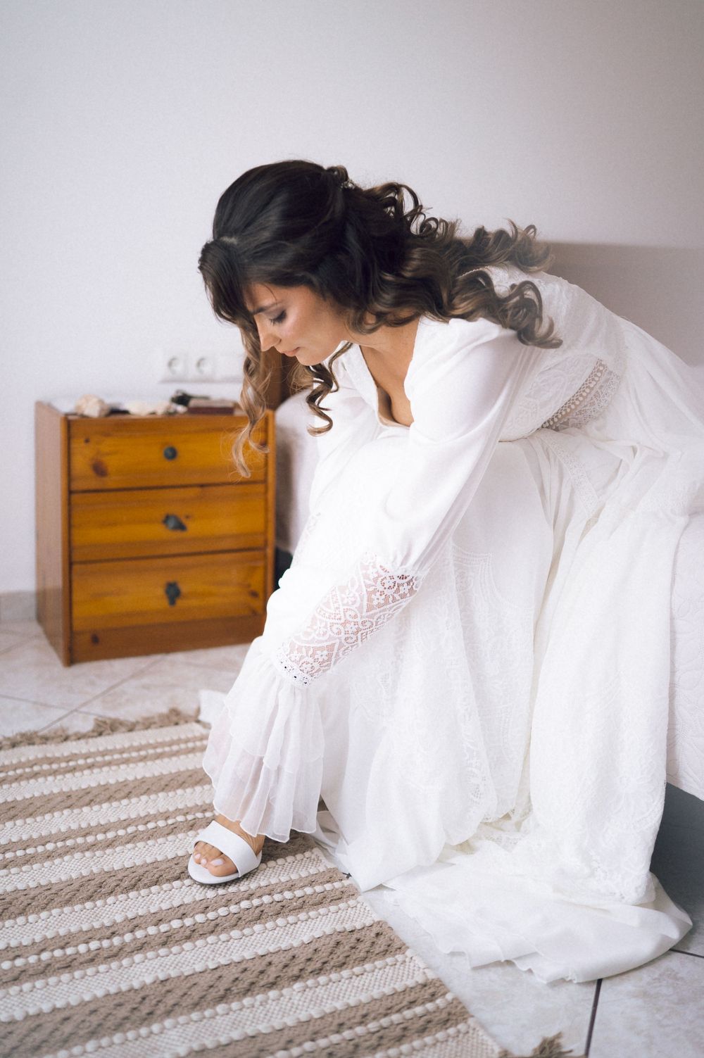 Bride in white wedding dress and lace sleeves sitting on bed putting on white sandals in bright bedroom.