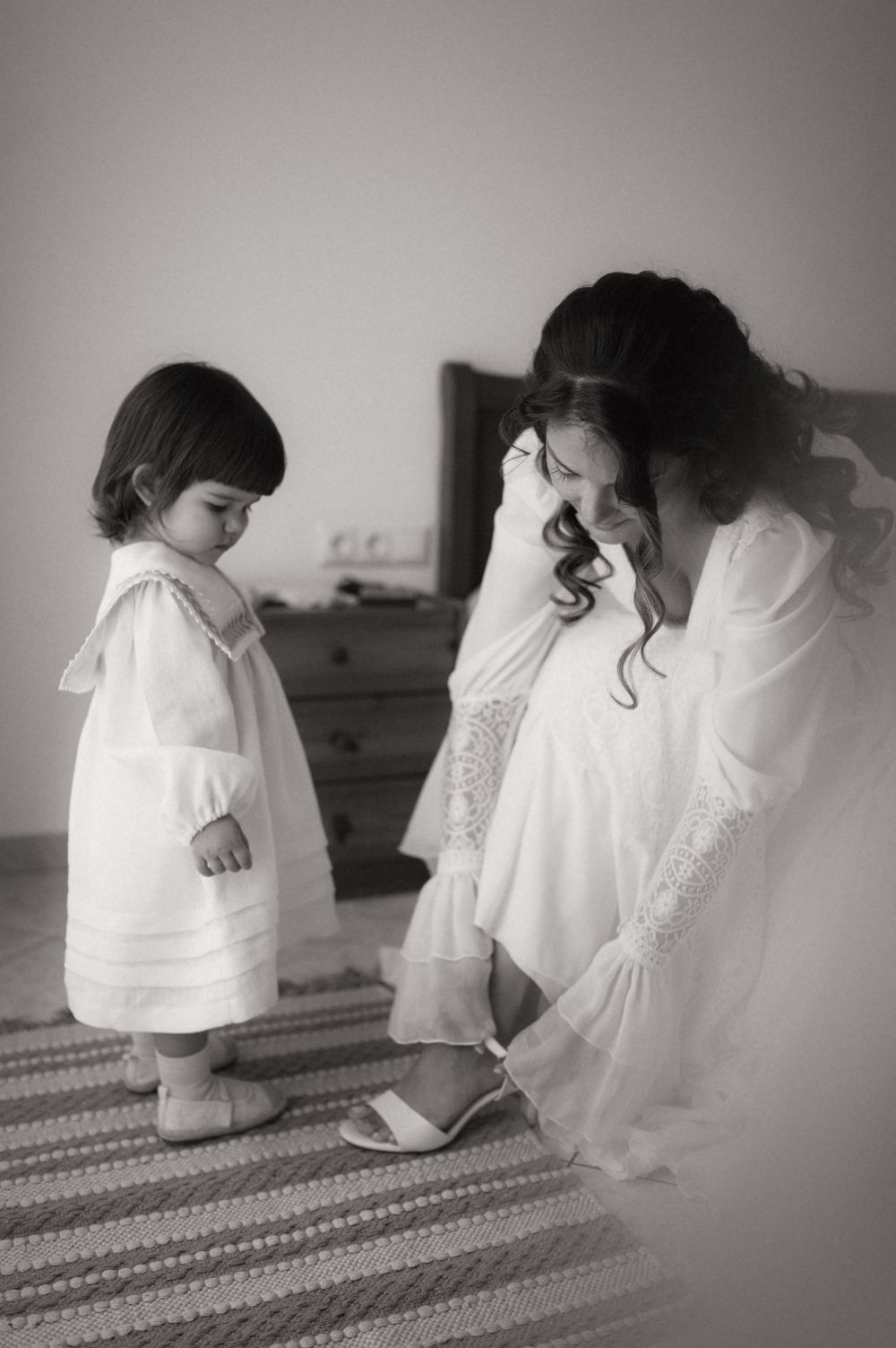 Black and white photo of a bride in white gown helping a toddler girl in white dress with shoes in a bedroom.