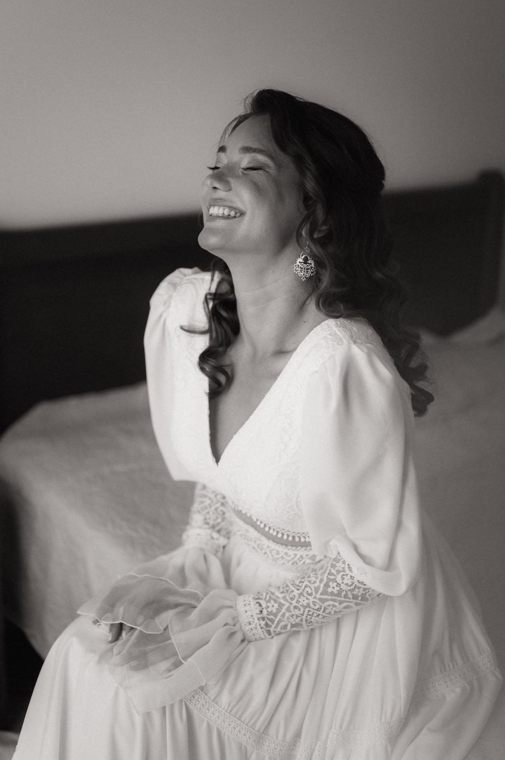Black and white photo of a smiling bride in a white lace dress sitting on a bed, laughing joyfully.