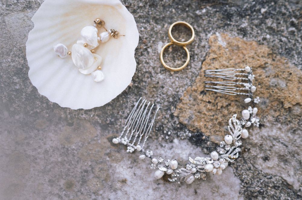 Bridal jewelry flatlay with pearl earrings, gold rings, crystal hair comb, and floral headpiece on stone surface.