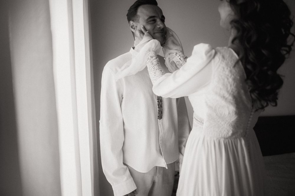 Black and white photo of a bride adjusting the groom's collar before their wedding ceremony.
