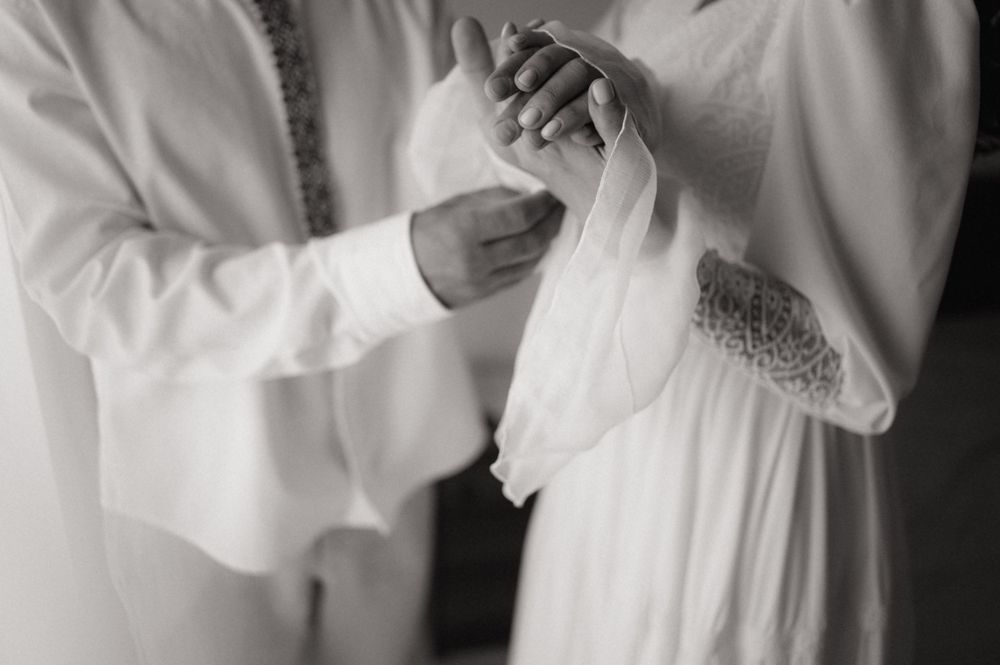 Black and white photo of two people in white garments holding hands during a wedding ceremony.