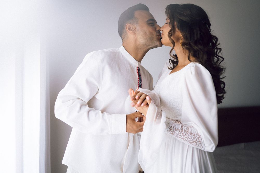 A couple in white attire shares a romantic kiss while holding hands in a bright, softly lit room.