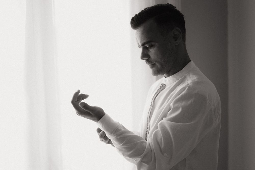Black and white photo of a man in a white dress shirt adjusting his cufflinks by a bright window.