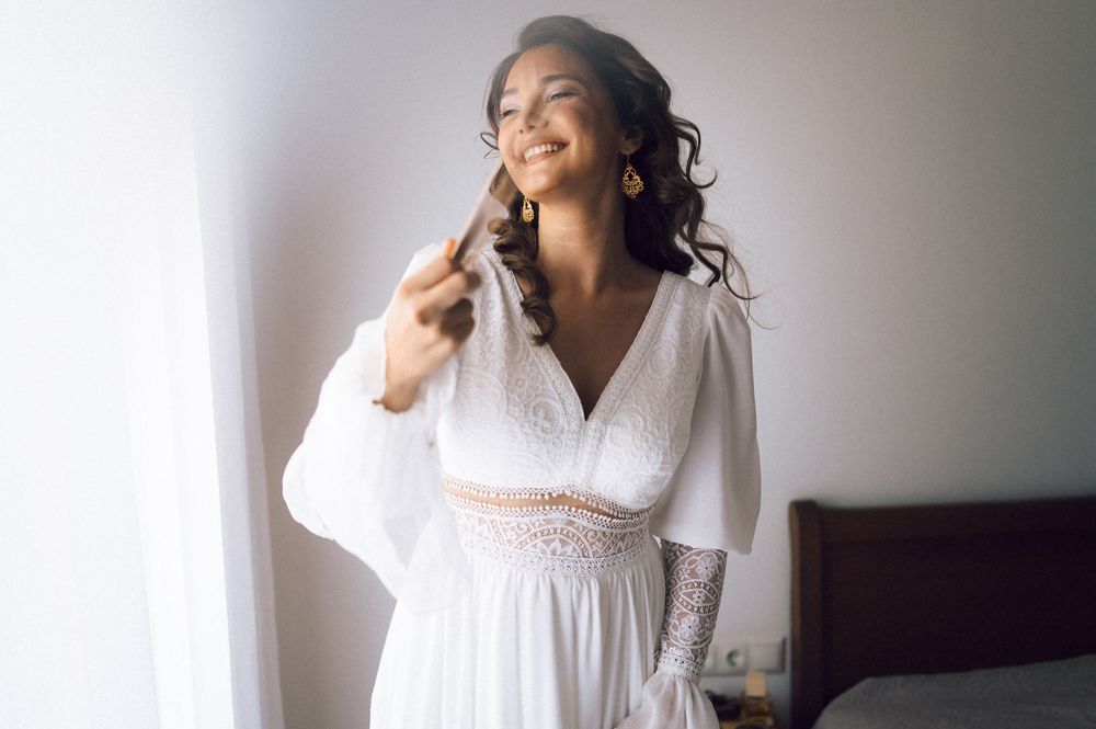 Smiling bride in a white boho wedding dress with lace details, posing by a bright window on her wedding day.