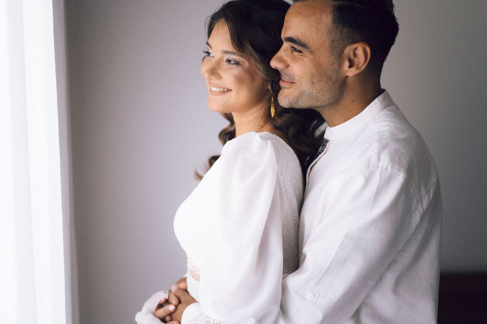 A happy couple in white outfits sharing a tender embrace near a bright window.
