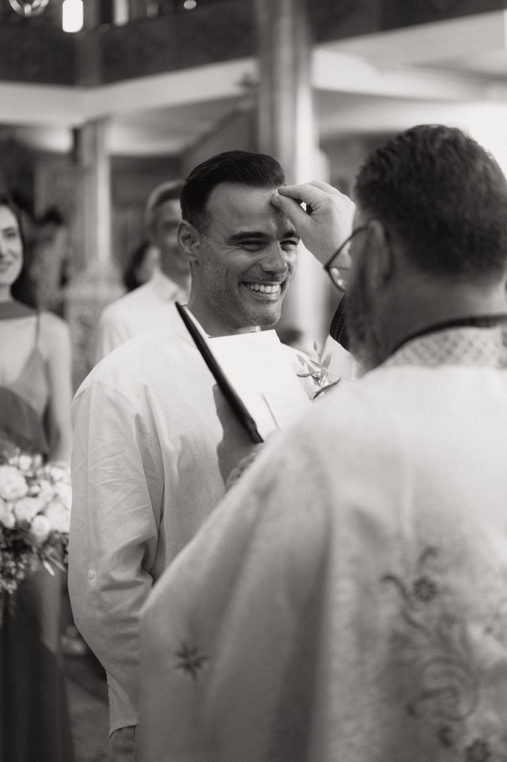 Black and white photo of a smiling groom receiving a blessing from a priest during a wedding ceremony.
