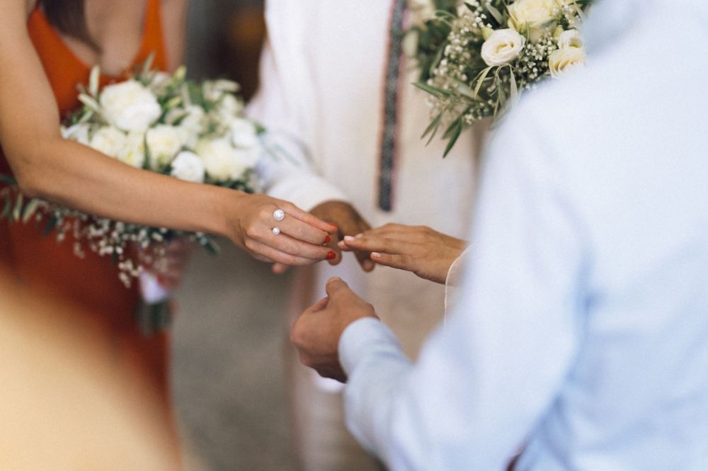 Couple exchanging wedding rings during ceremony, bride holding white floral bouquet with greenery.