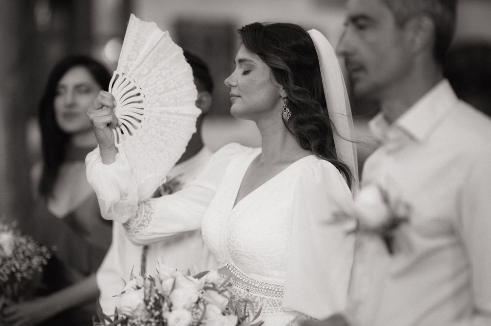 Elegant bride in white gown holding a decorative fan and bouquet at a wedding ceremony, black and white photo.