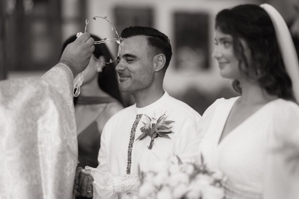 Black and white wedding ceremony photo of groom smiling as officiant performs blessing with bride holding bouquet.