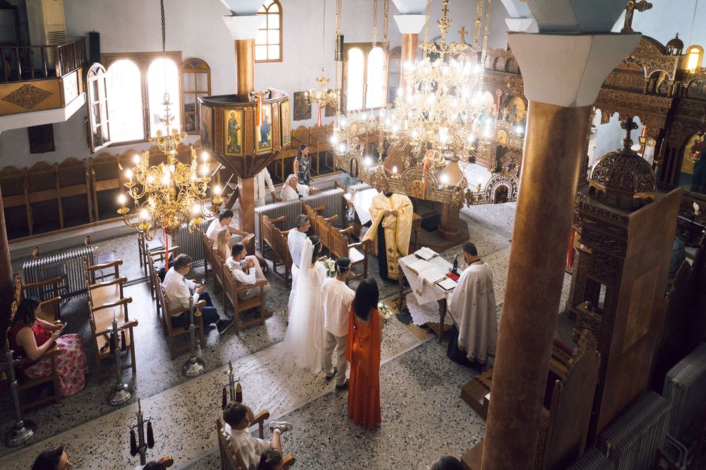 Orthodox church ceremony with clergy and congregation gathered under ornate chandeliers in a decorated sanctuary.