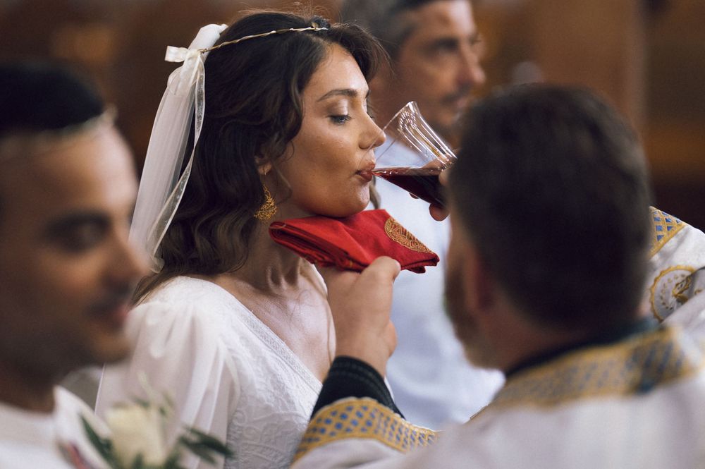 Bride in white dress and veil drinking red wine from a chalice during a religious wedding ceremony.
