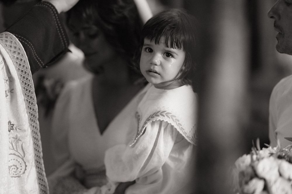 Black and white photo of a young girl in white dress being held at a wedding ceremony.