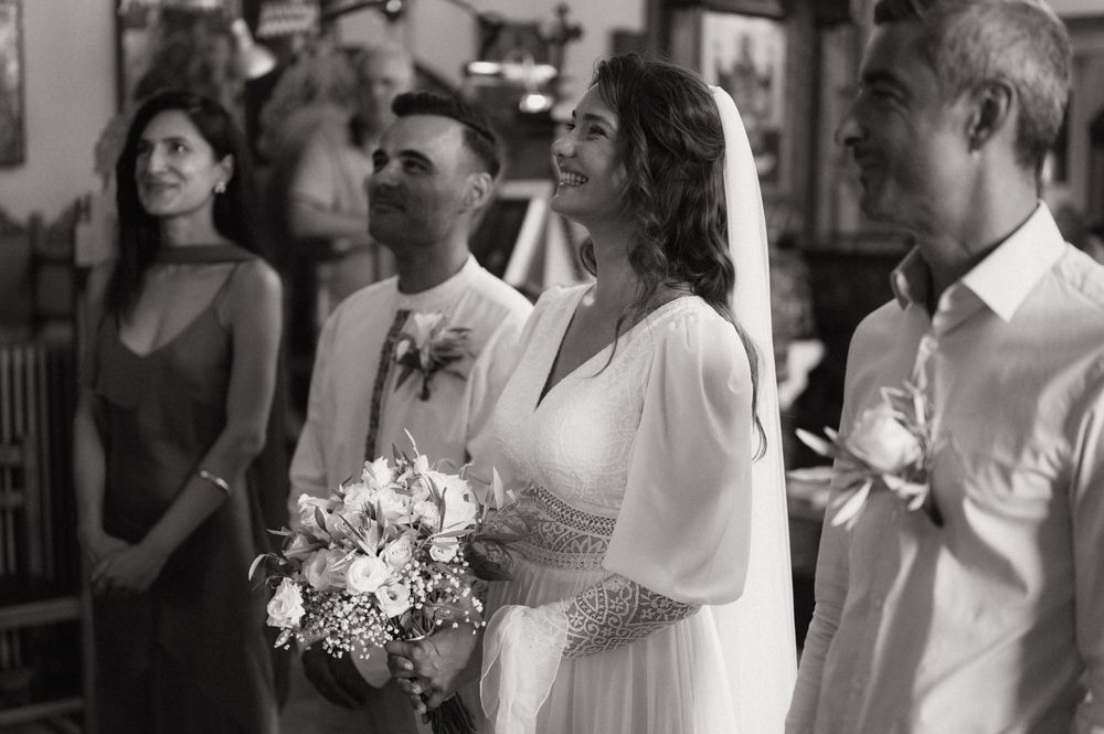 Black and white wedding photo of bride holding bouquet with groom and guests smiling during ceremony.
