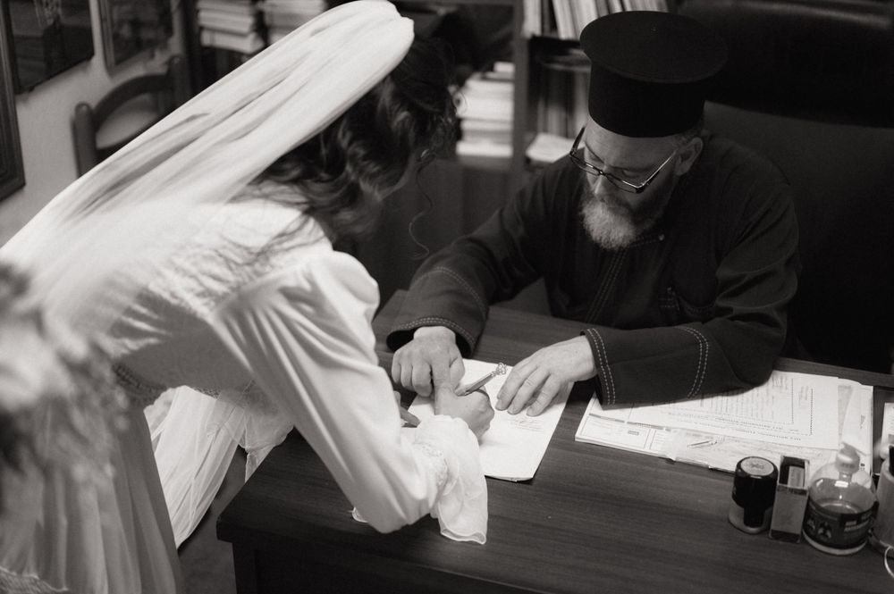 Black and white photo of a bride in white veil signing documents with an orthodox priest at a desk.