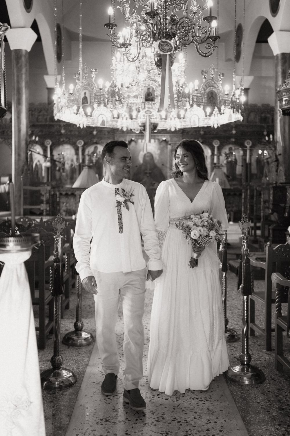 Black and white photo of a couple in wedding attire walking hand in hand down a church aisle under a chandelier.
