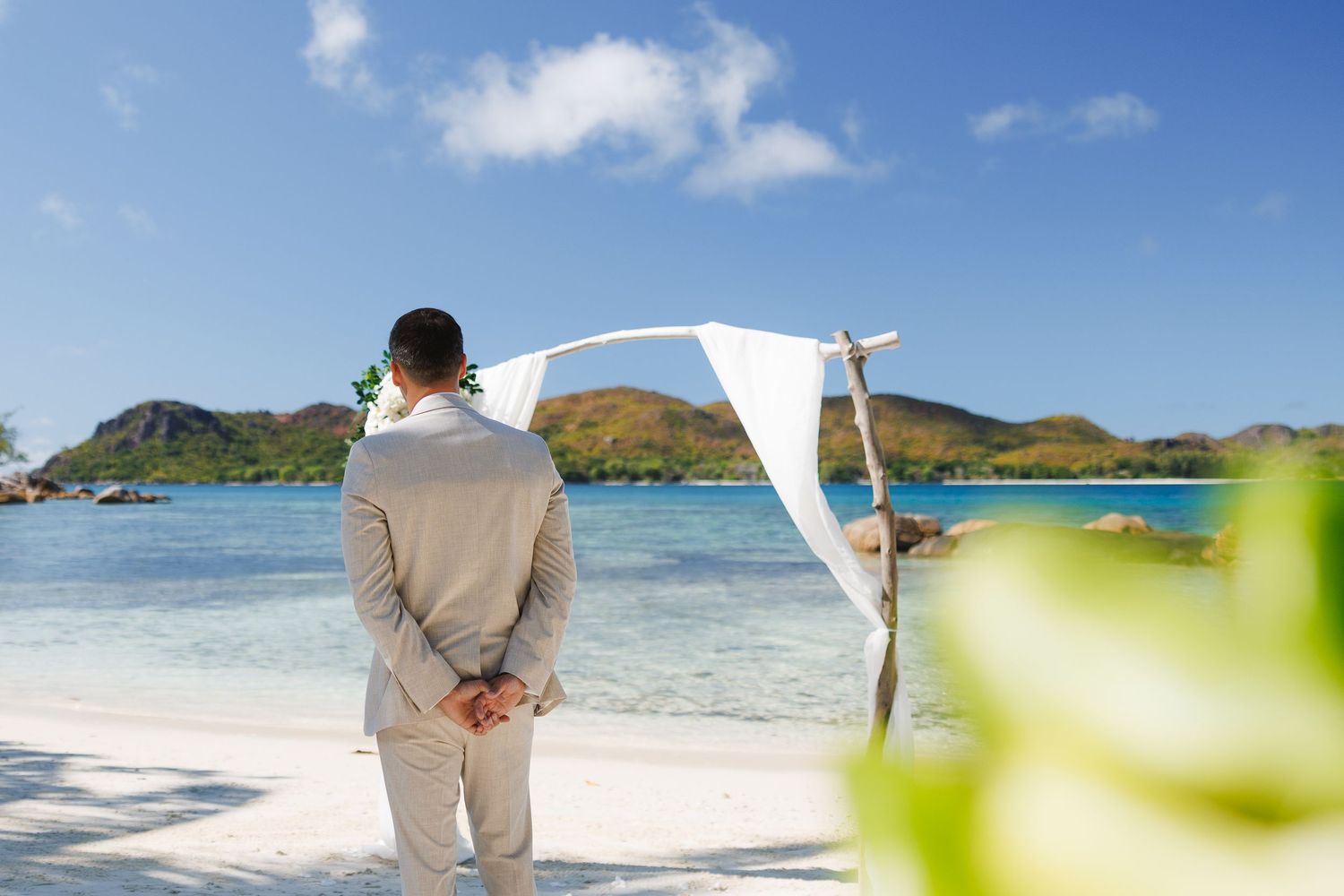 Groom in a gray suit standing at a tropical beach wedding altar with white draping and ocean views.