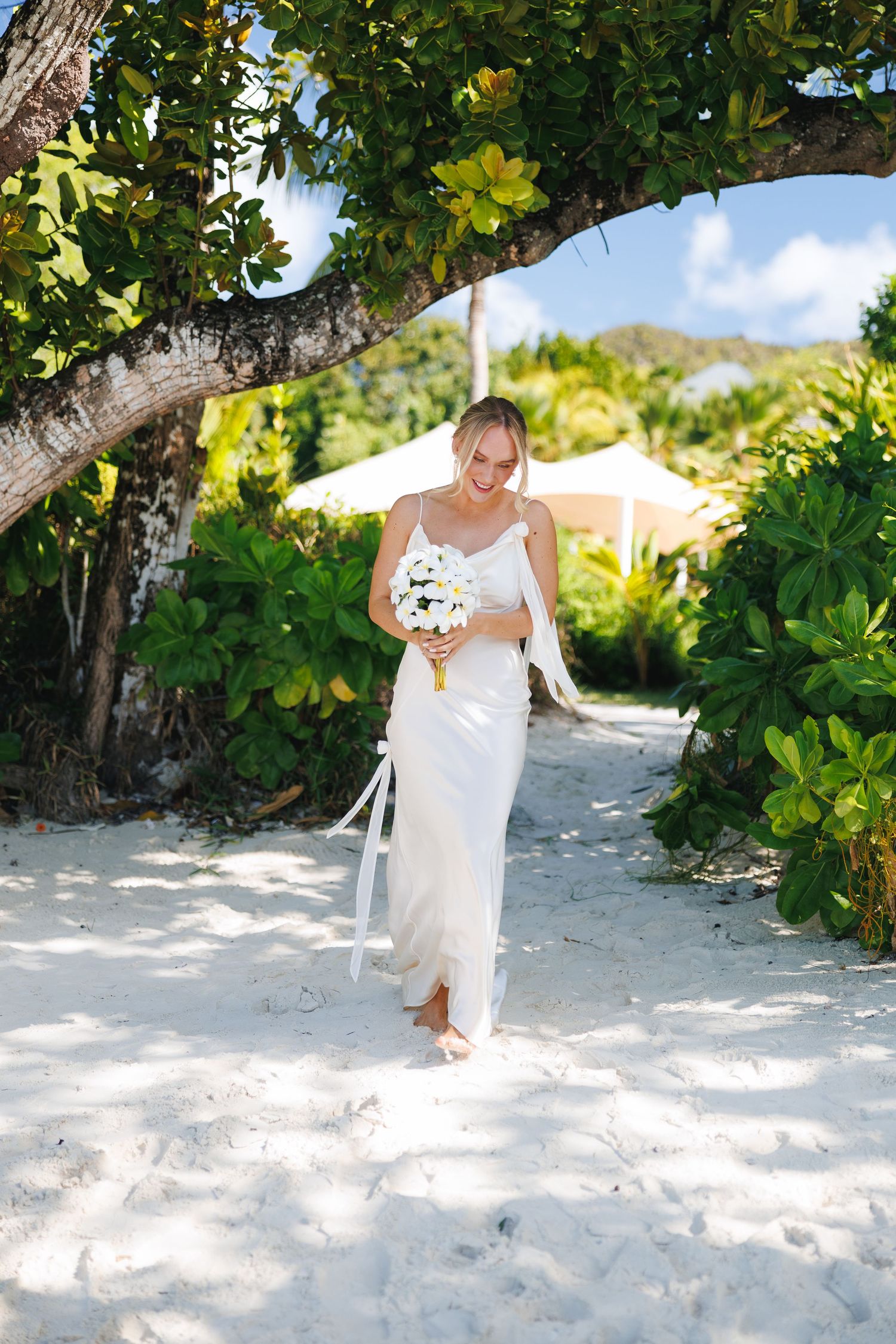 Bride in a white slip dress walking barefoot on a sandy beach holding a white flower bouquet.