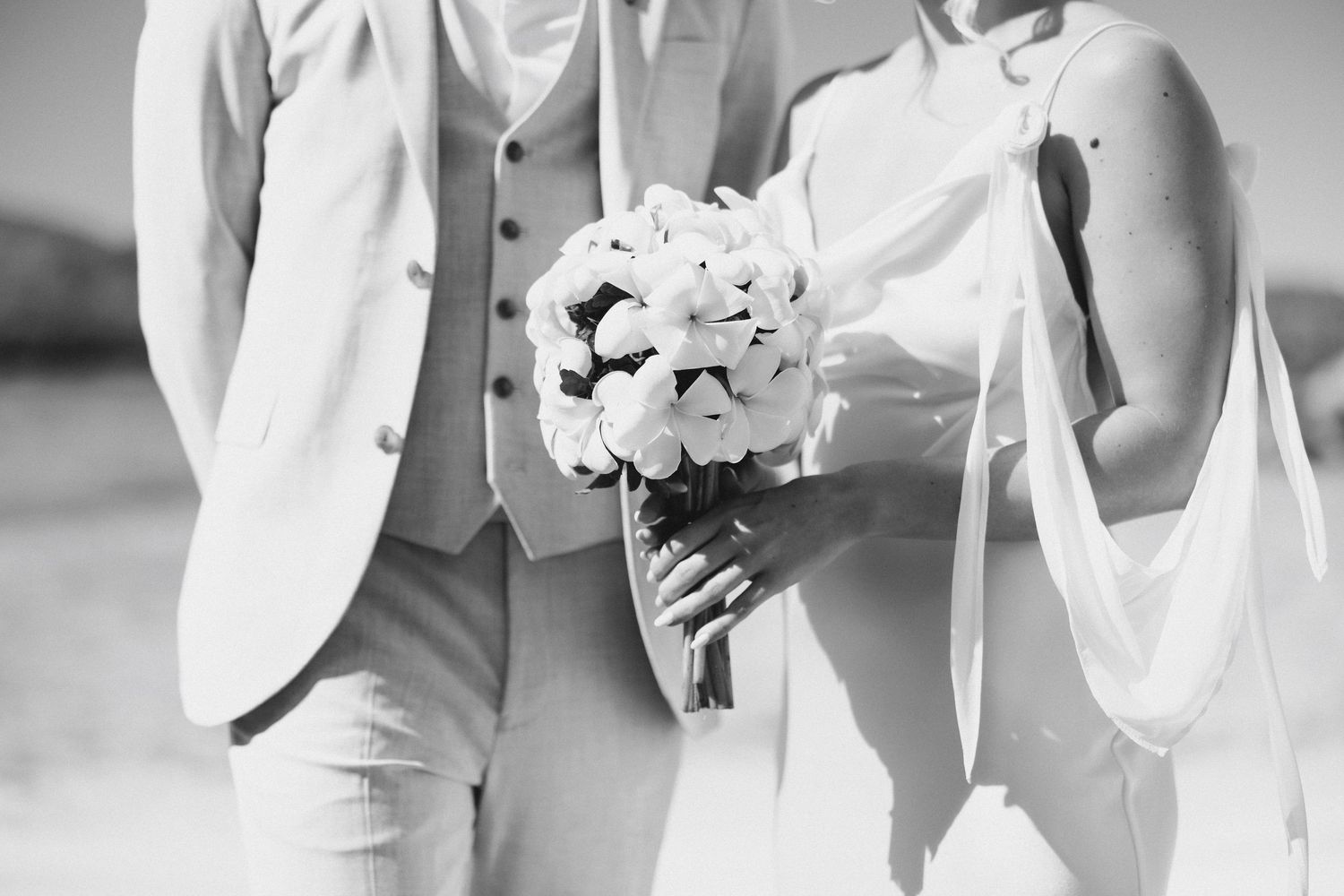 Black and white photo of bride and groom holding a white floral bouquet on a sunny beach.