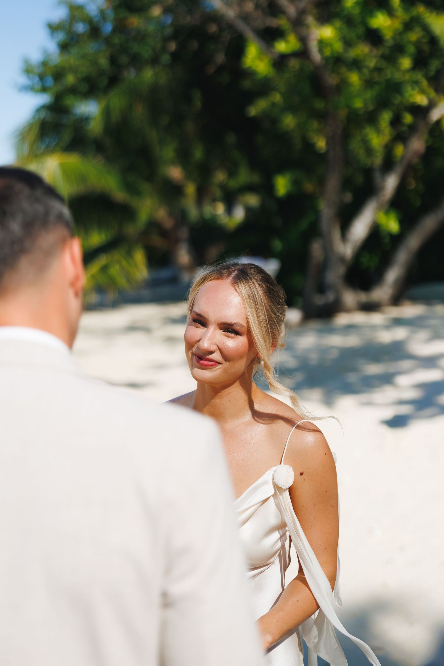 Smiling bride in a white satin dress gazing at her groom during a tropical beach wedding.