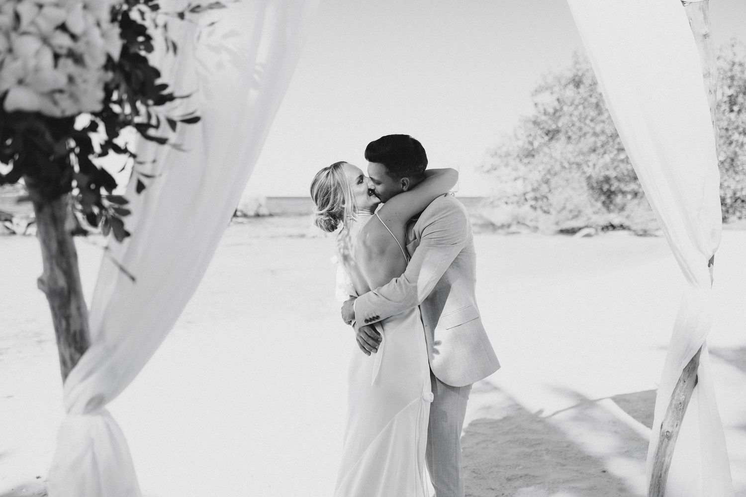 Black and white photo of bride and groom sharing their first kiss under a draped beach arch.