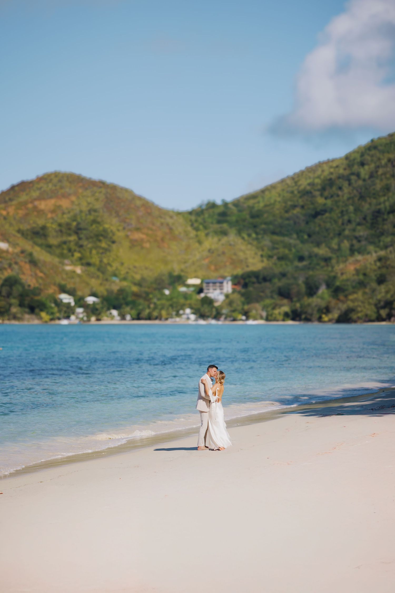 Bride and groom embracing on a white sand beach with turquoise water and lush green hills in the background.