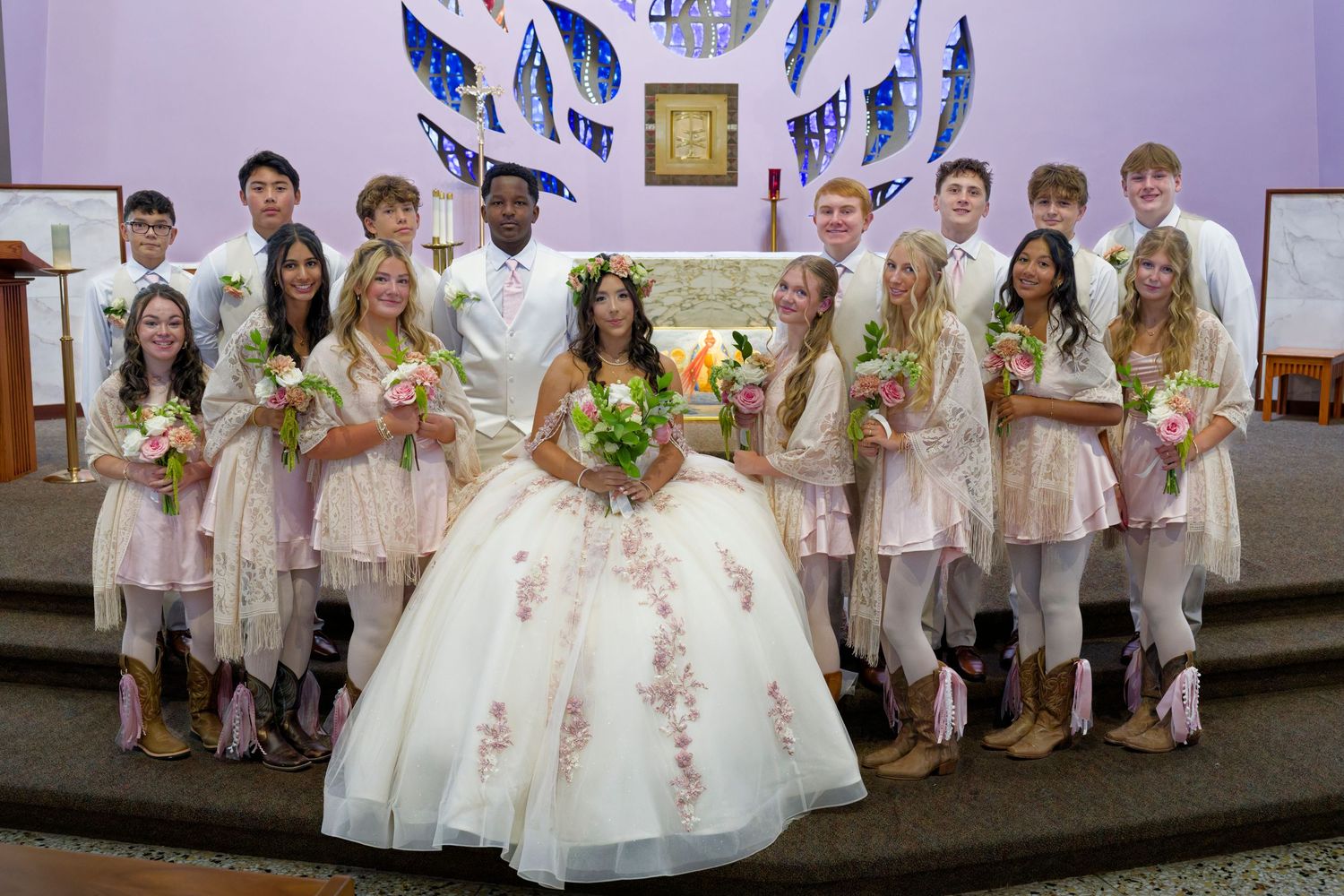 Quinceañera court poses at church altar with girl in white floral gown surrounded by chambelanes and damas.