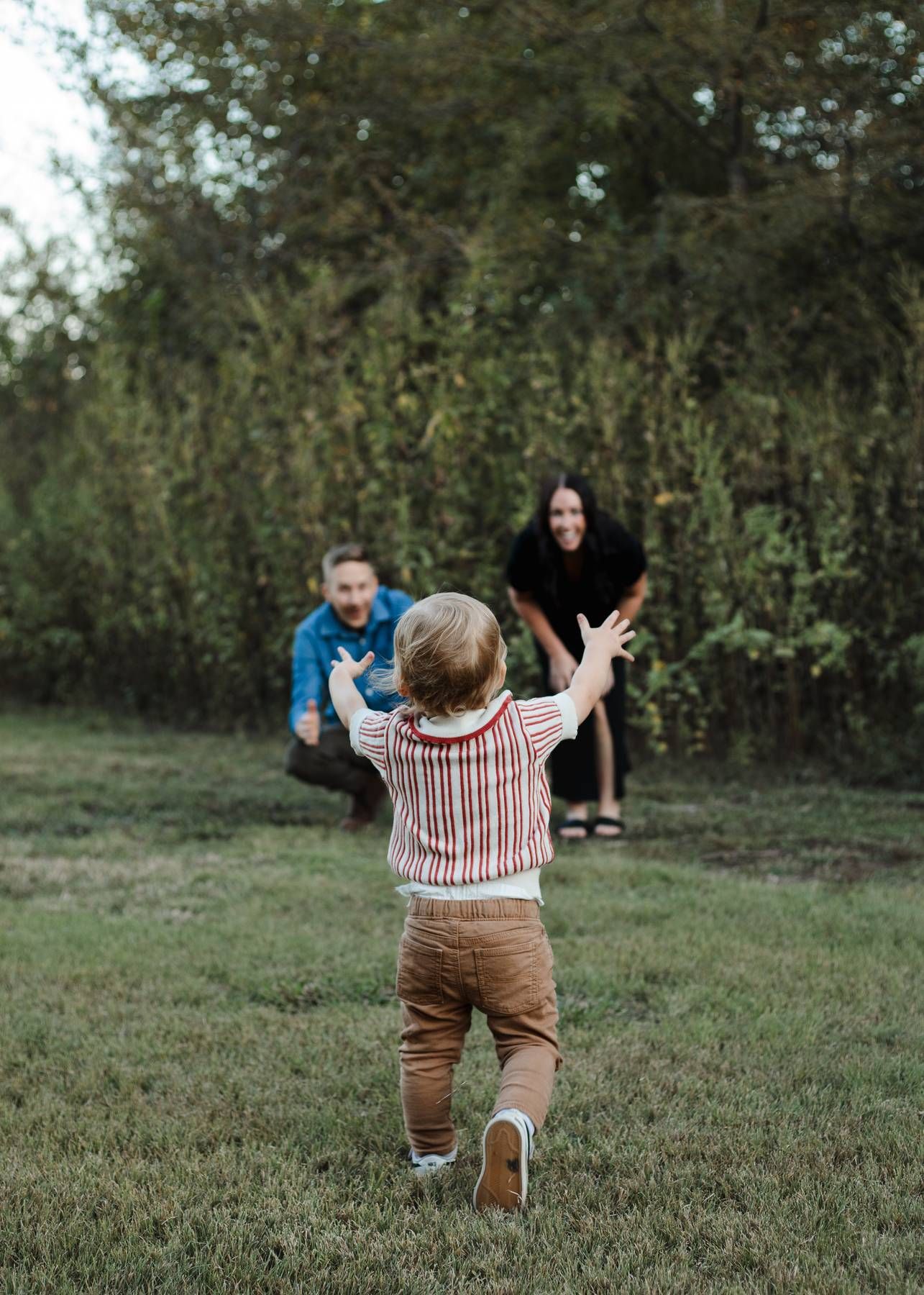 Toddler in a striped shirt runs across a grassy lawn toward his smiling parents crouching with open arms outdoors.