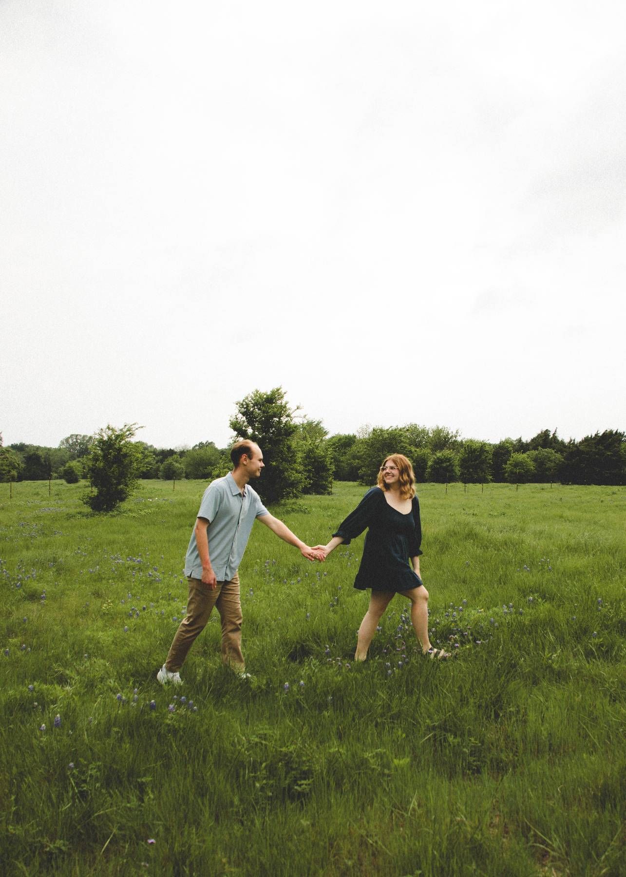 Engaged couple holds hands and walks through a lush green meadow under a soft overcast sky.