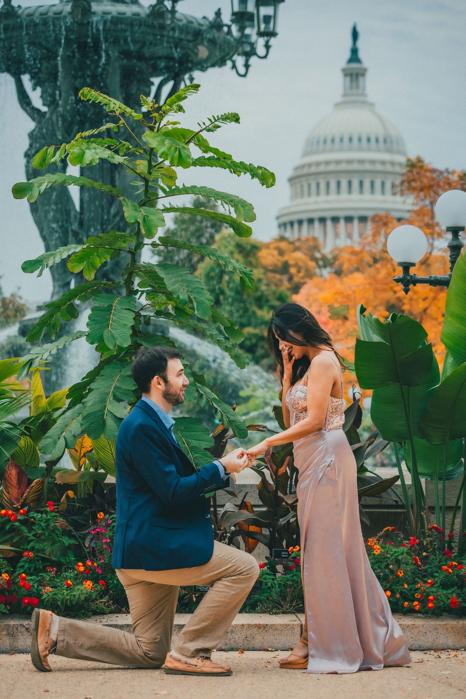 Man in a navy blazer kneels on one knee proposing to a smiling woman in a blush gown near autumn foliage with the U.S. Capitol dome behind.