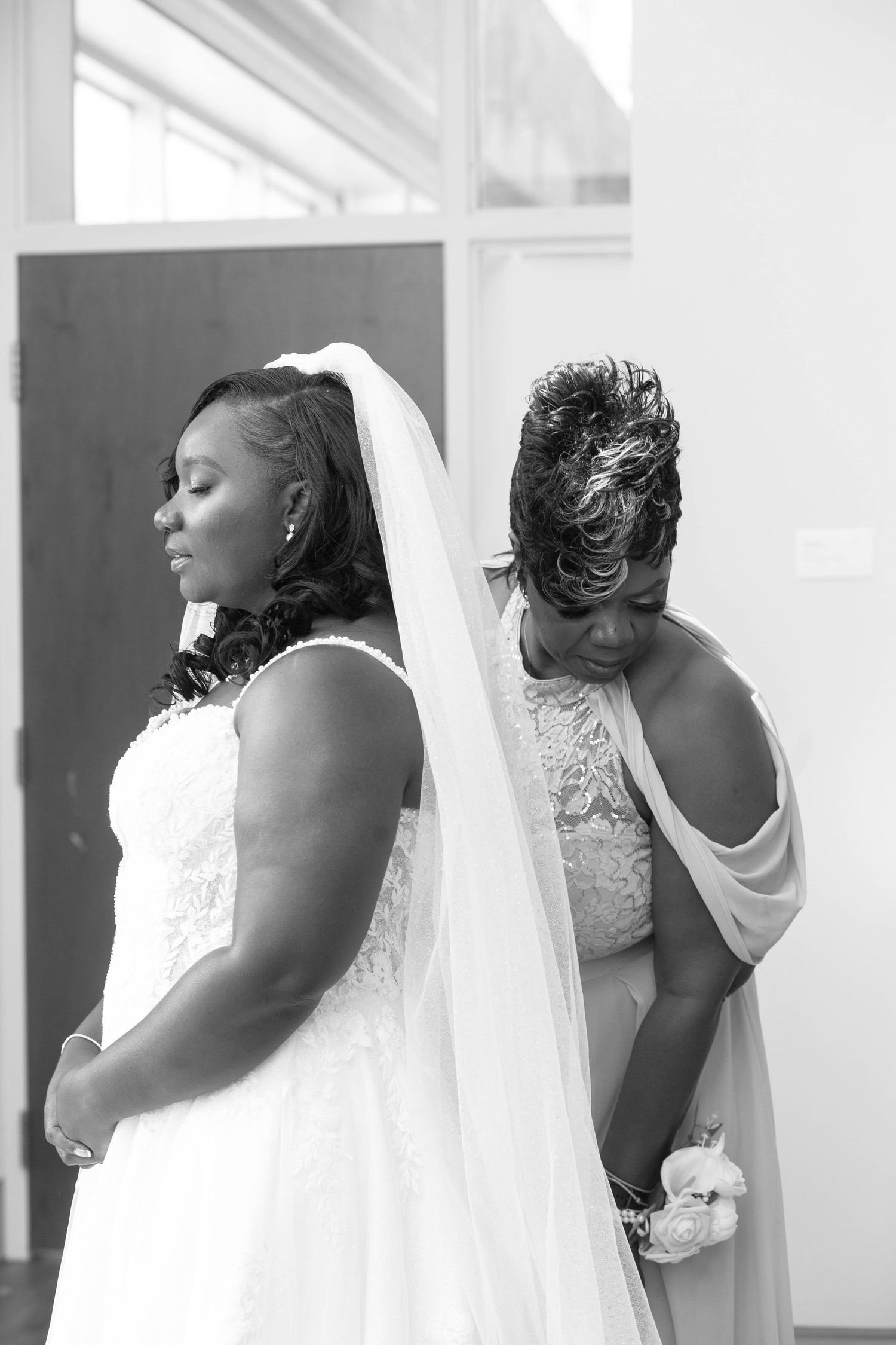 Black and white photo of a bride in a lace gown and veil standing back to back with a woman in a draped dress holding a floral corsage.