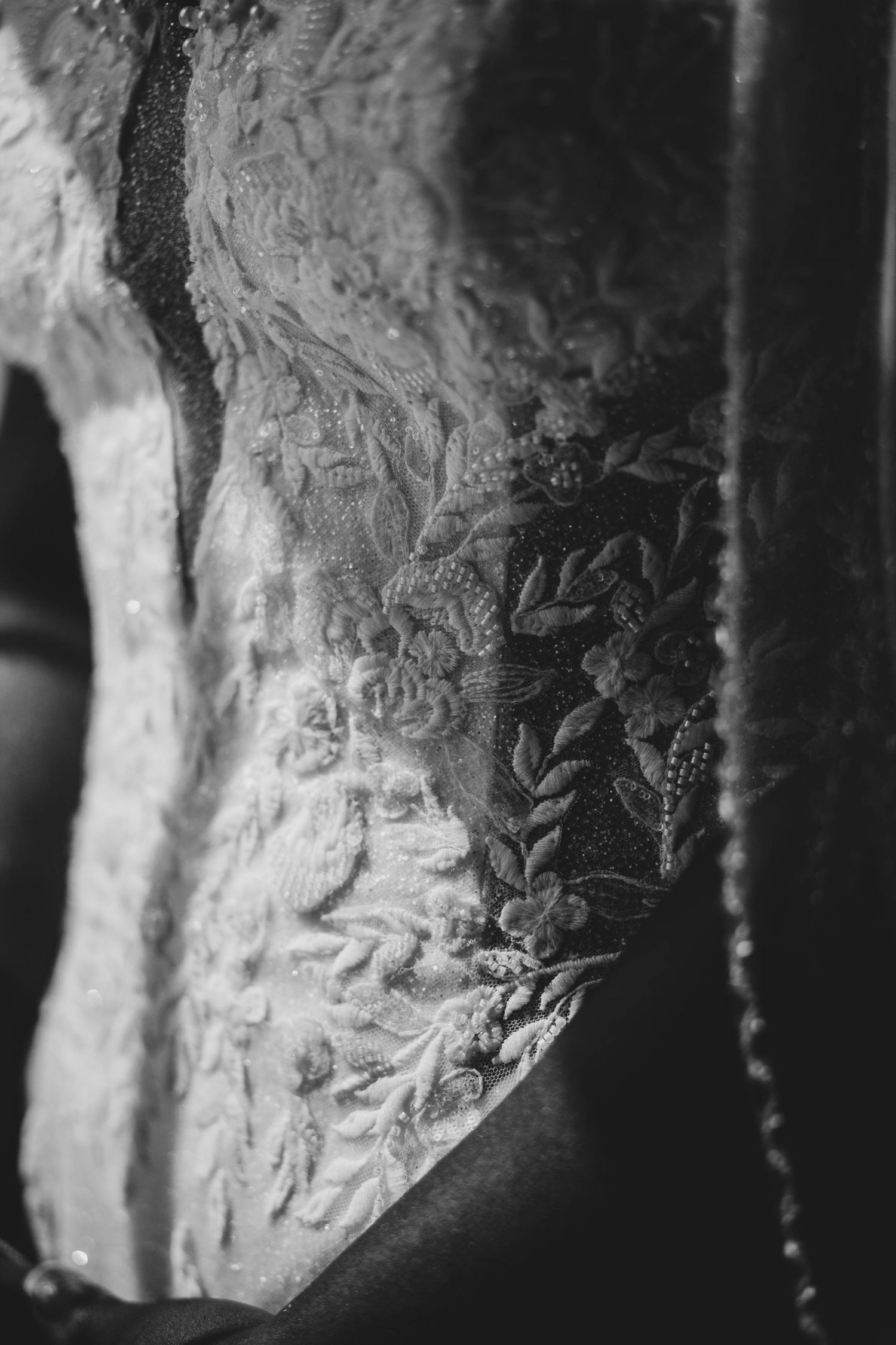 Black and white close-up of an intricate lace wedding gown bodice with floral embroidery and delicate beaded details.